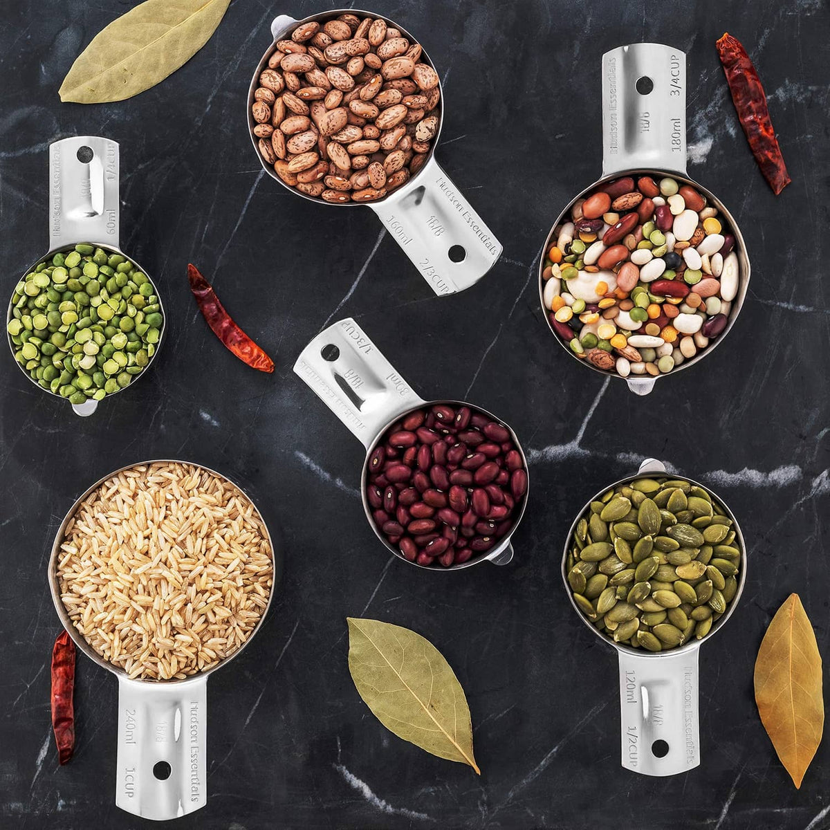 Six stainless steel measuring cups filled with assorted beans and seeds