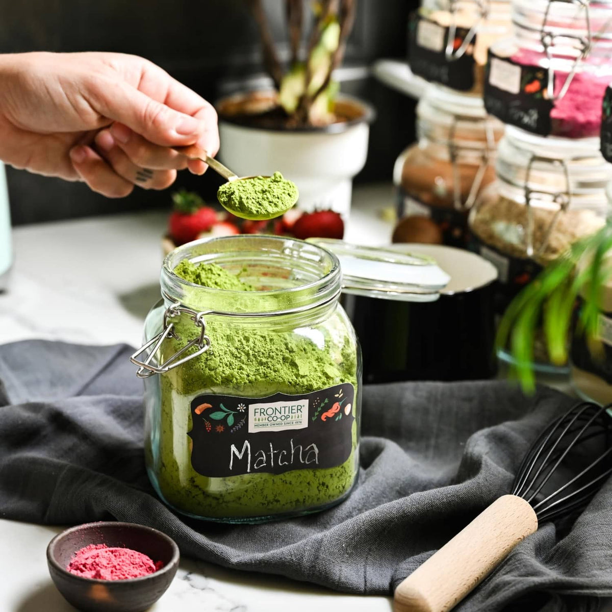 Open glass jar of green matcha tea powder with wooden scoop and whisk