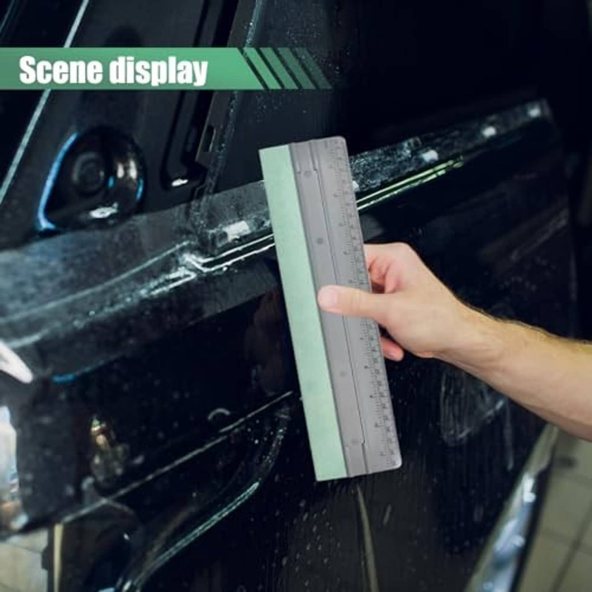 Vinyl wrap squeegee tool with gray ruler and mint pad, held against vehicle glass