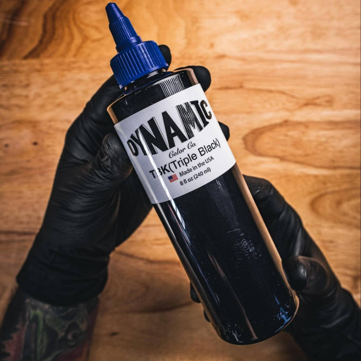 Dynamic triple black ink bottle in blue cap held in black-gloved hand.