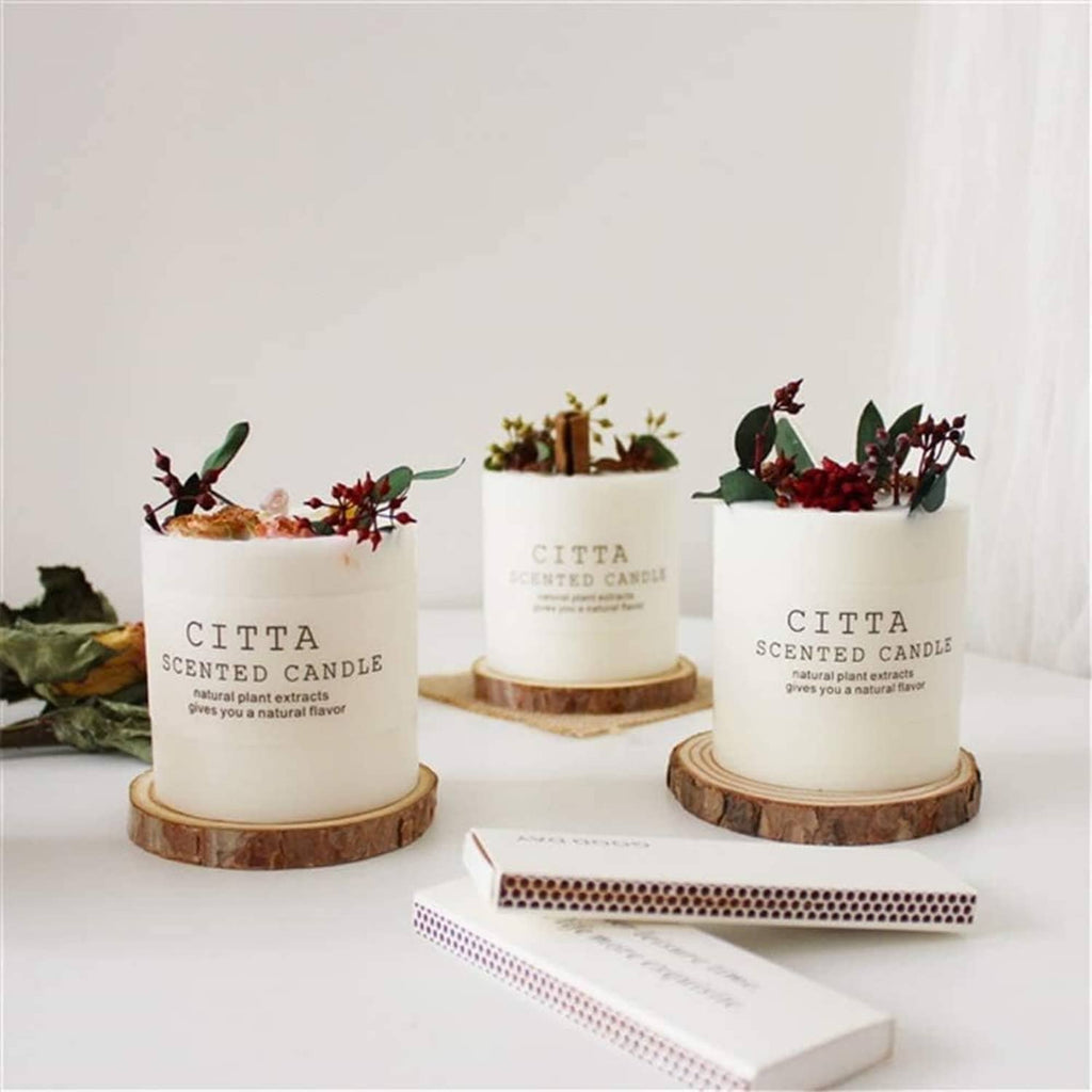 Set of white cylindrical candles with dried botanicals placed in paper labels on wood bases.
