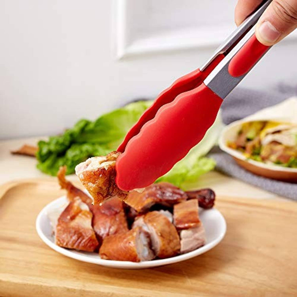 Red silicone kitchen tongs lifting meat from a bowl on a wooden table.