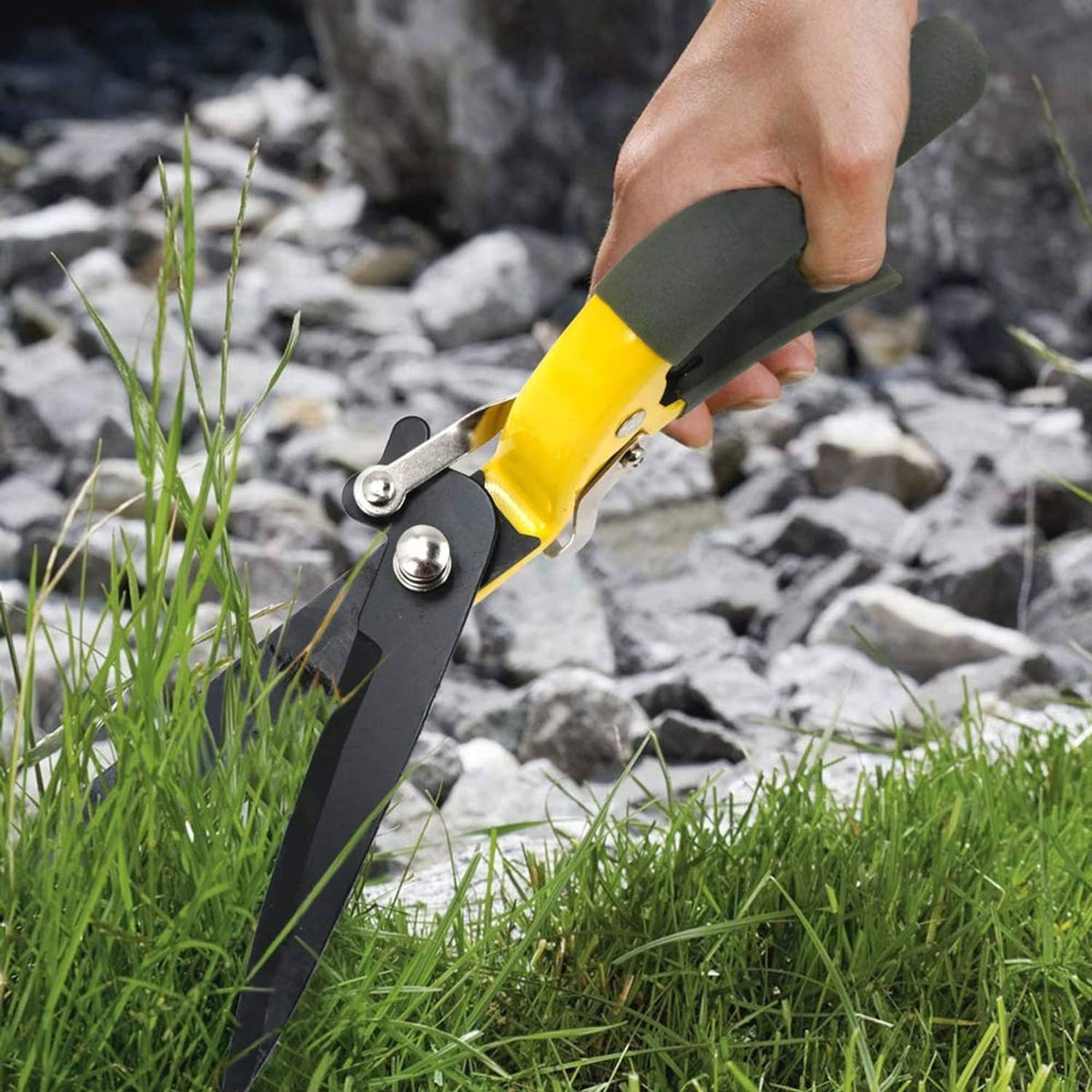 Hand holding yellow grass shears with gray blades, trimming grass along rocky ground.
