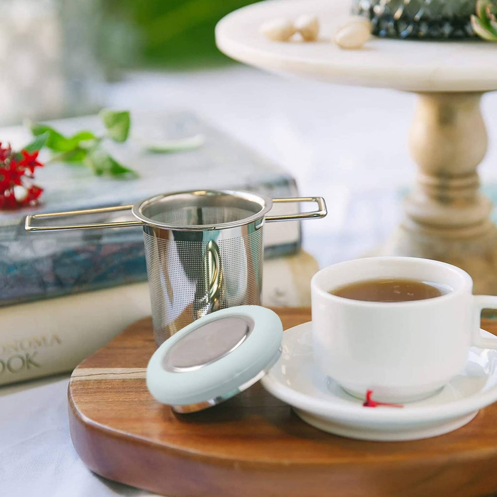 Stainless tea infuser with light blue lid on a tray beside a cup of tea