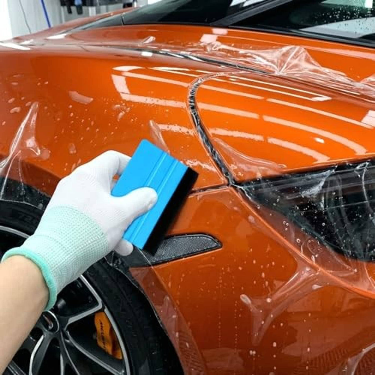 Blue felt-edge squeegee pressed on car window film by gloved hand.