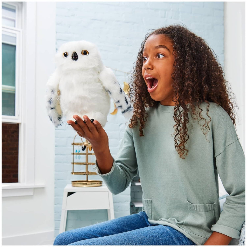 White interactive owl plush held up by a person, showing amber eyes and spotted wings.