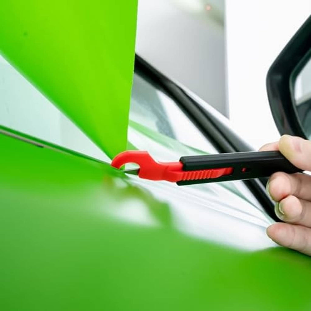 Hand using red-and-black utility knife to trim neon green window tint film