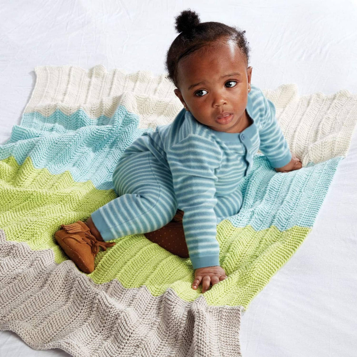 Baby sitting on a striped knit blanket in light blue, green, and cream