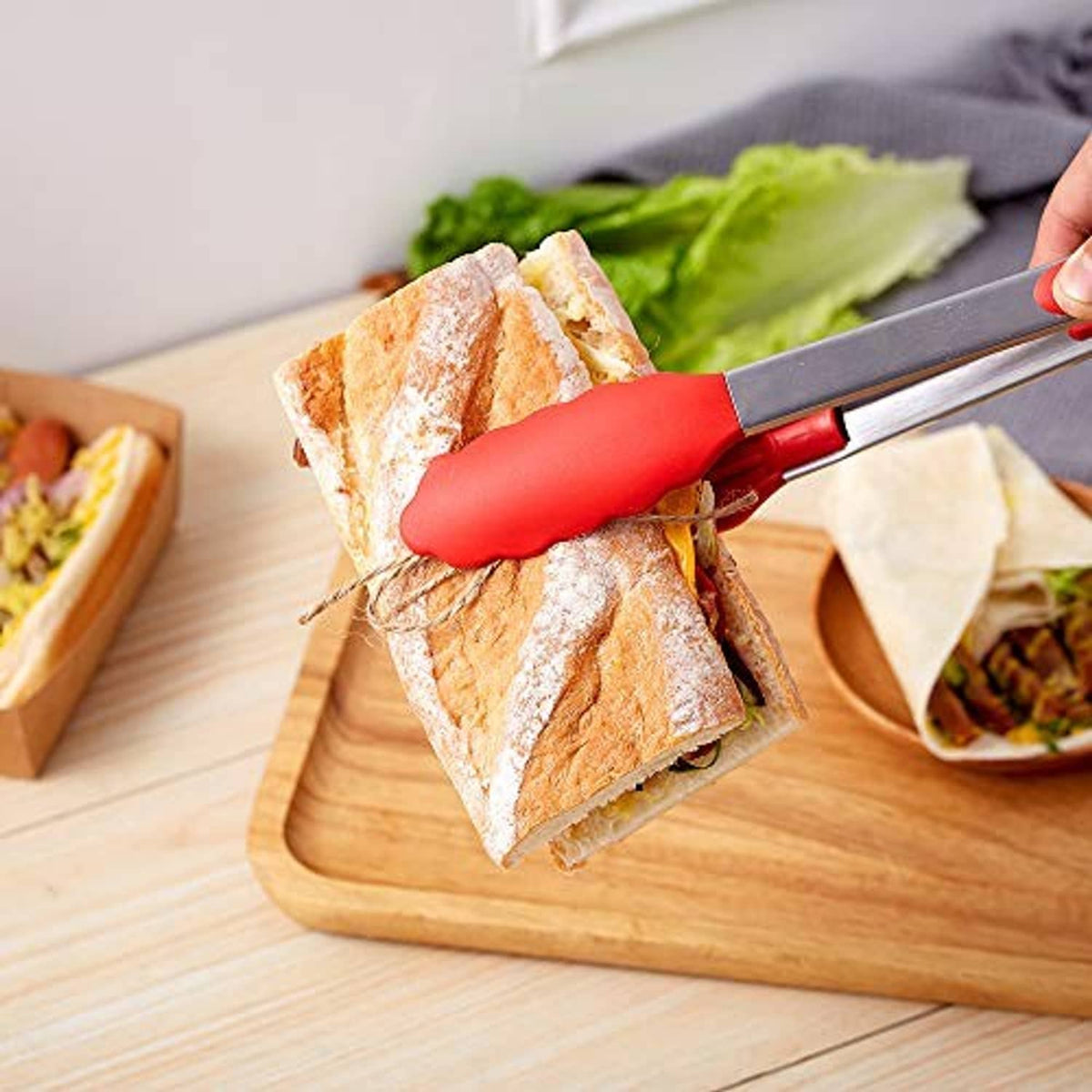 Red-handled stainless steel kitchen tongs lifting sandwiches over a wooden board.