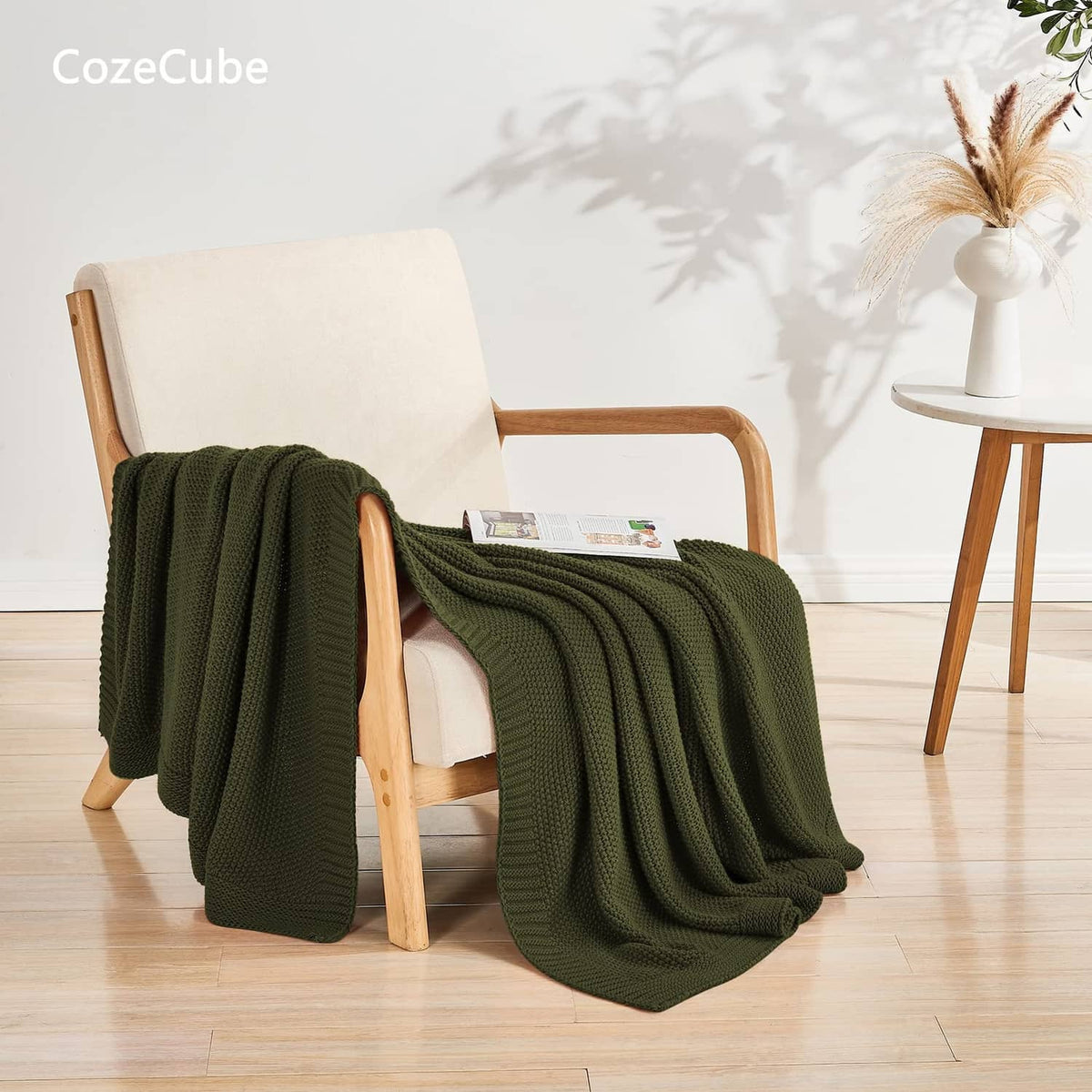 Olive green knit throw blanket spread over a wooden armchair and floor.