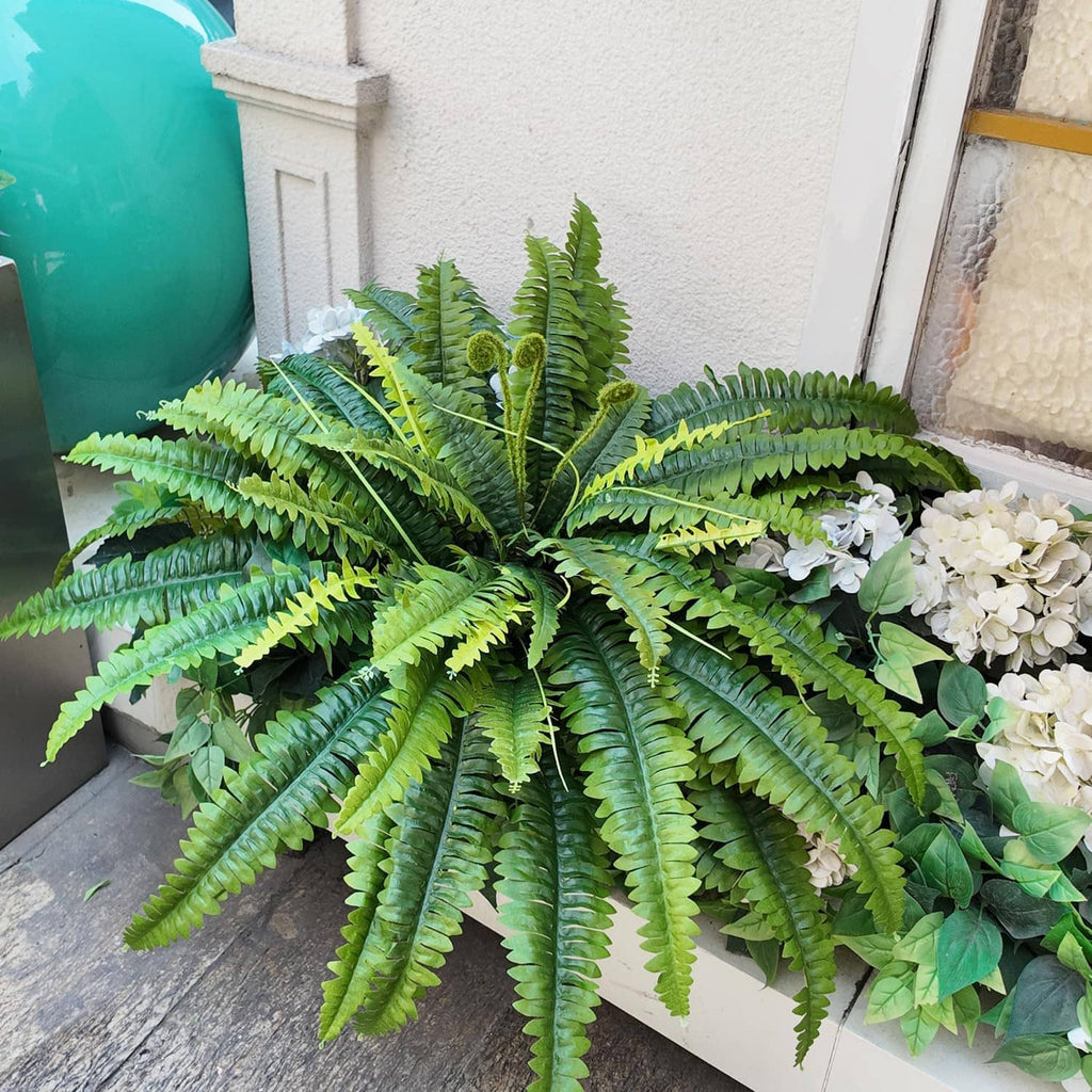 Set of 2 artificial green ferns with wide fronds, styled beside white flowers.