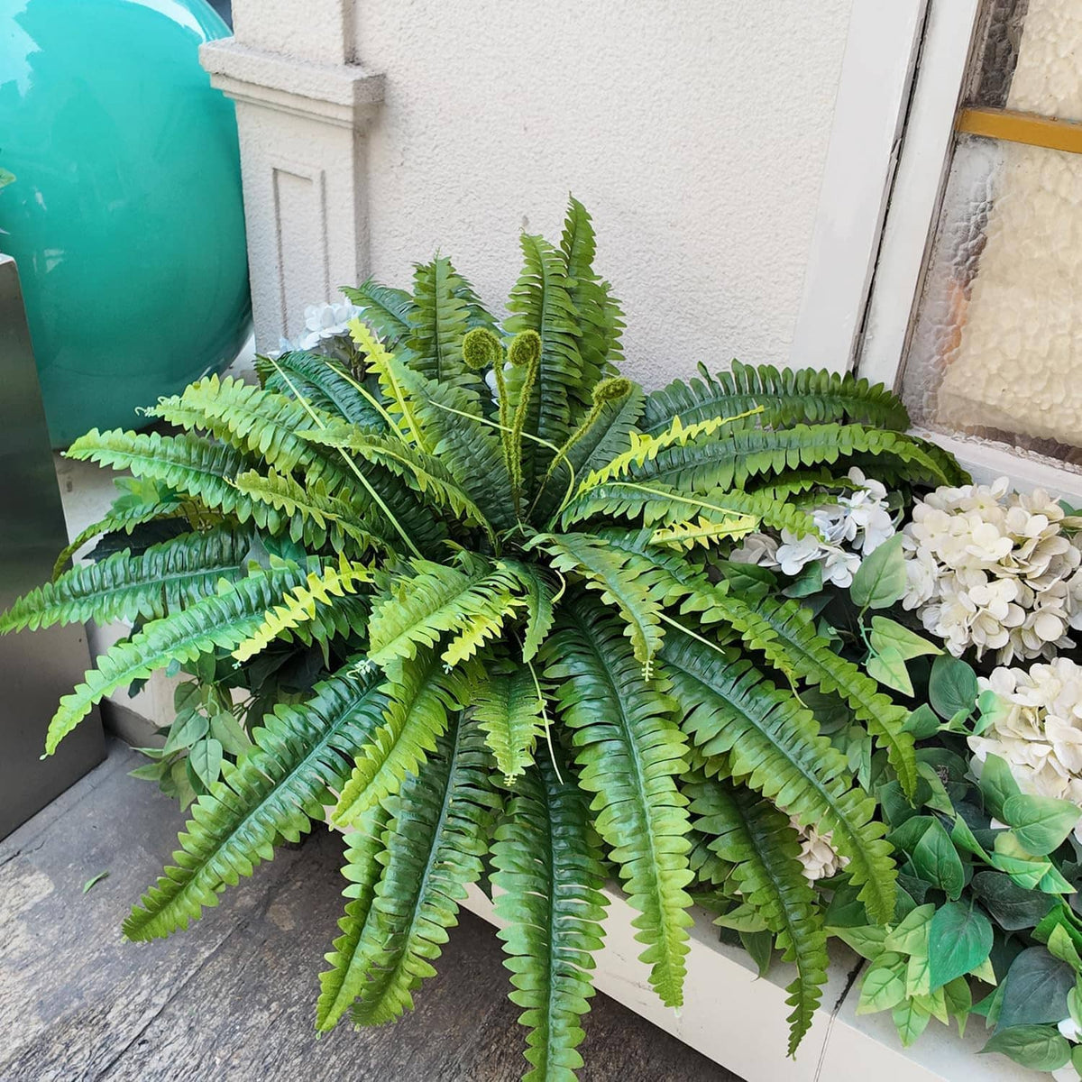 Set of 2 artificial green ferns with wide fronds, styled beside white flowers.