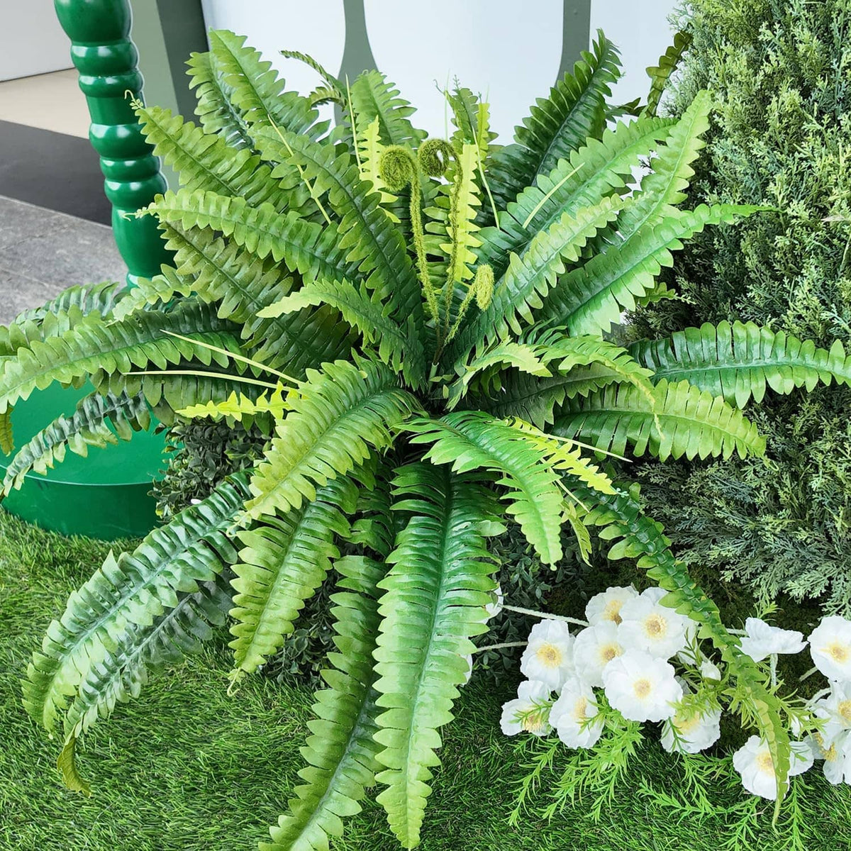 Set of 2 artificial green ferns with dense fronds, positioned among hedges and grass.