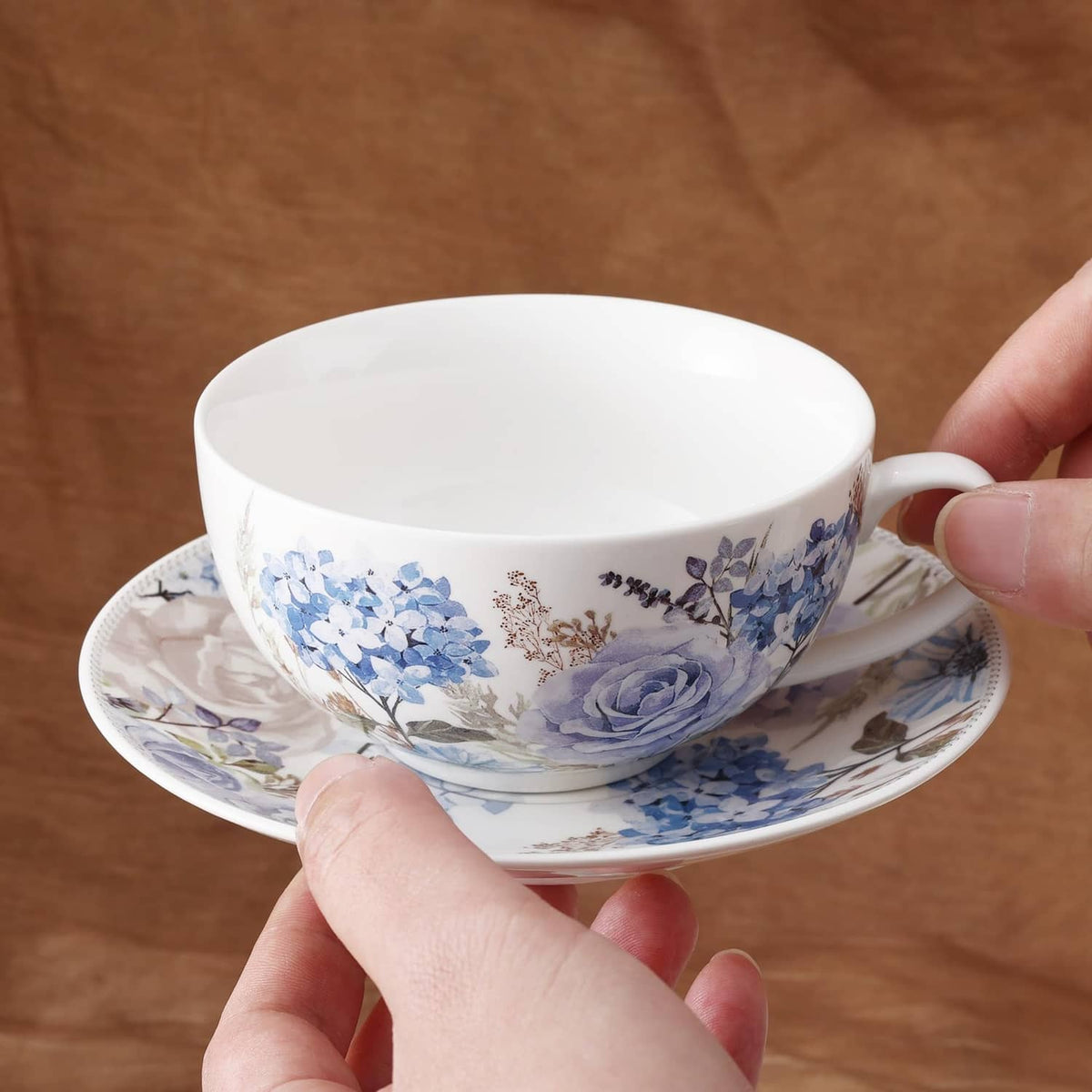Porcelain teacup and saucer with blue floral rose pattern, shown being held.