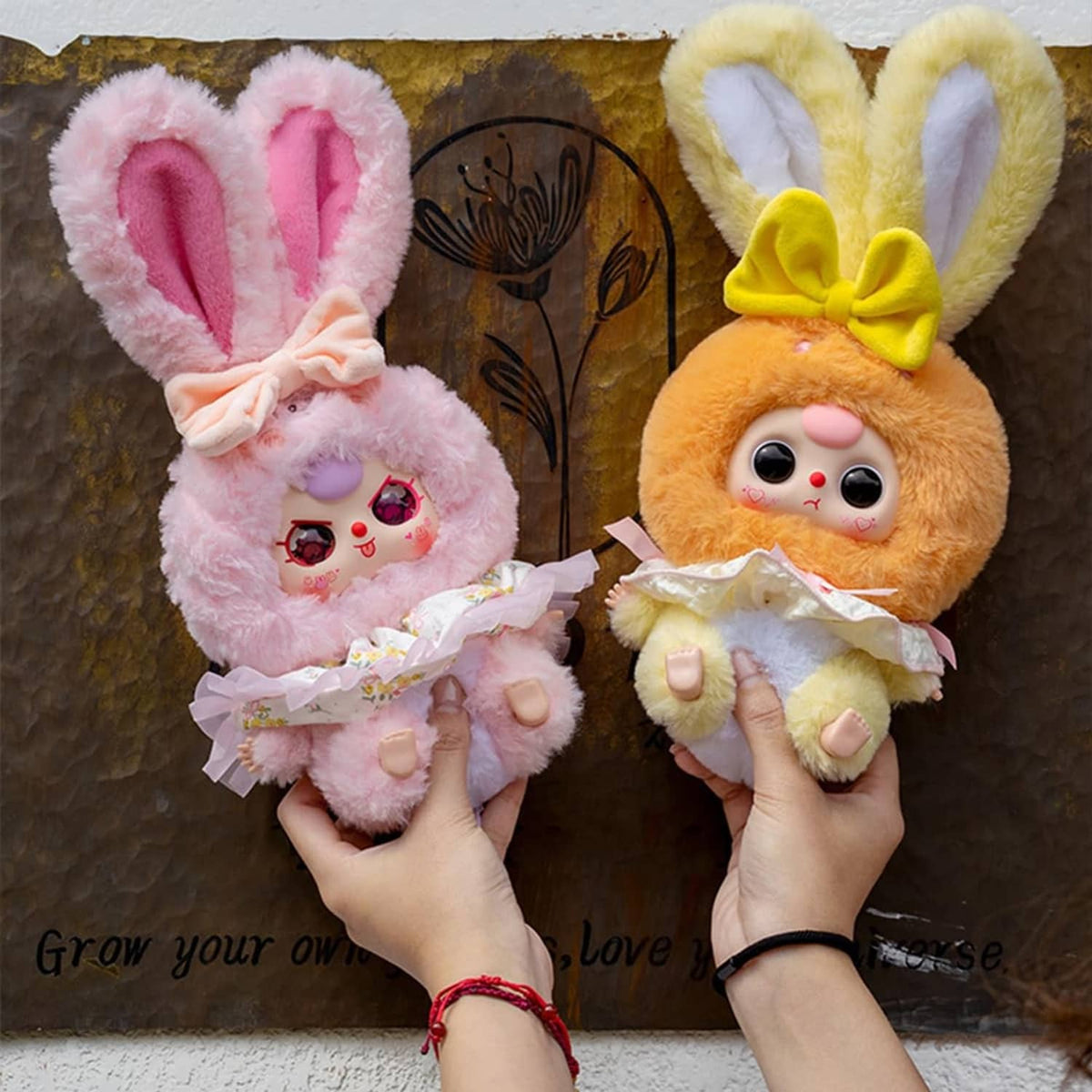 Pink and yellow-orange rabbit plush figures held in hands, each with ruffled outfit