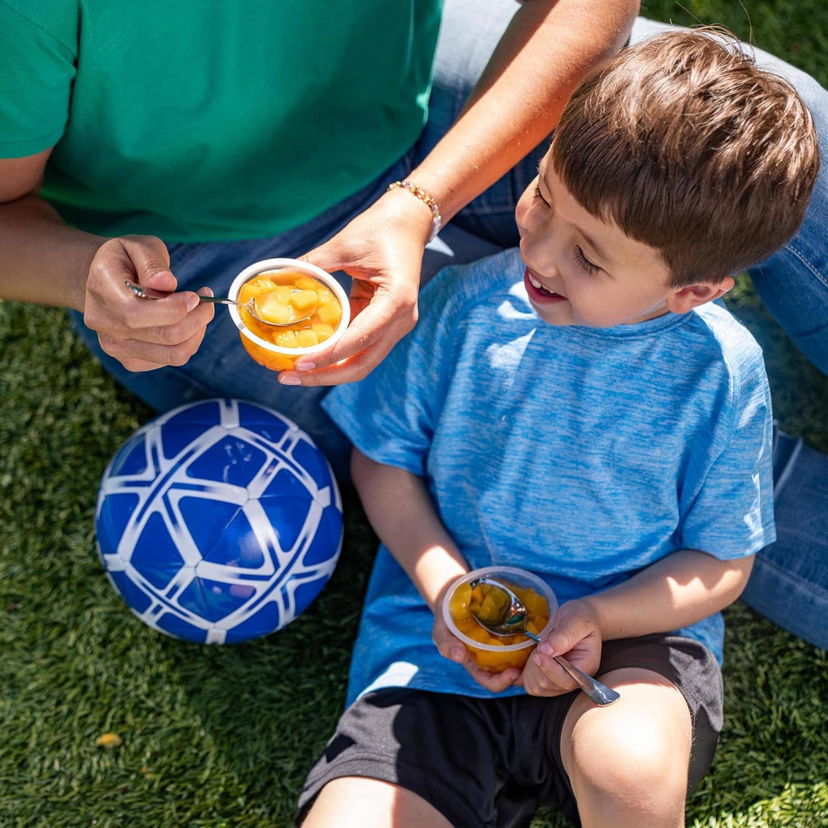 Boy eating fruit from a cup with a spoon while seated outdoors, with snacks nearby