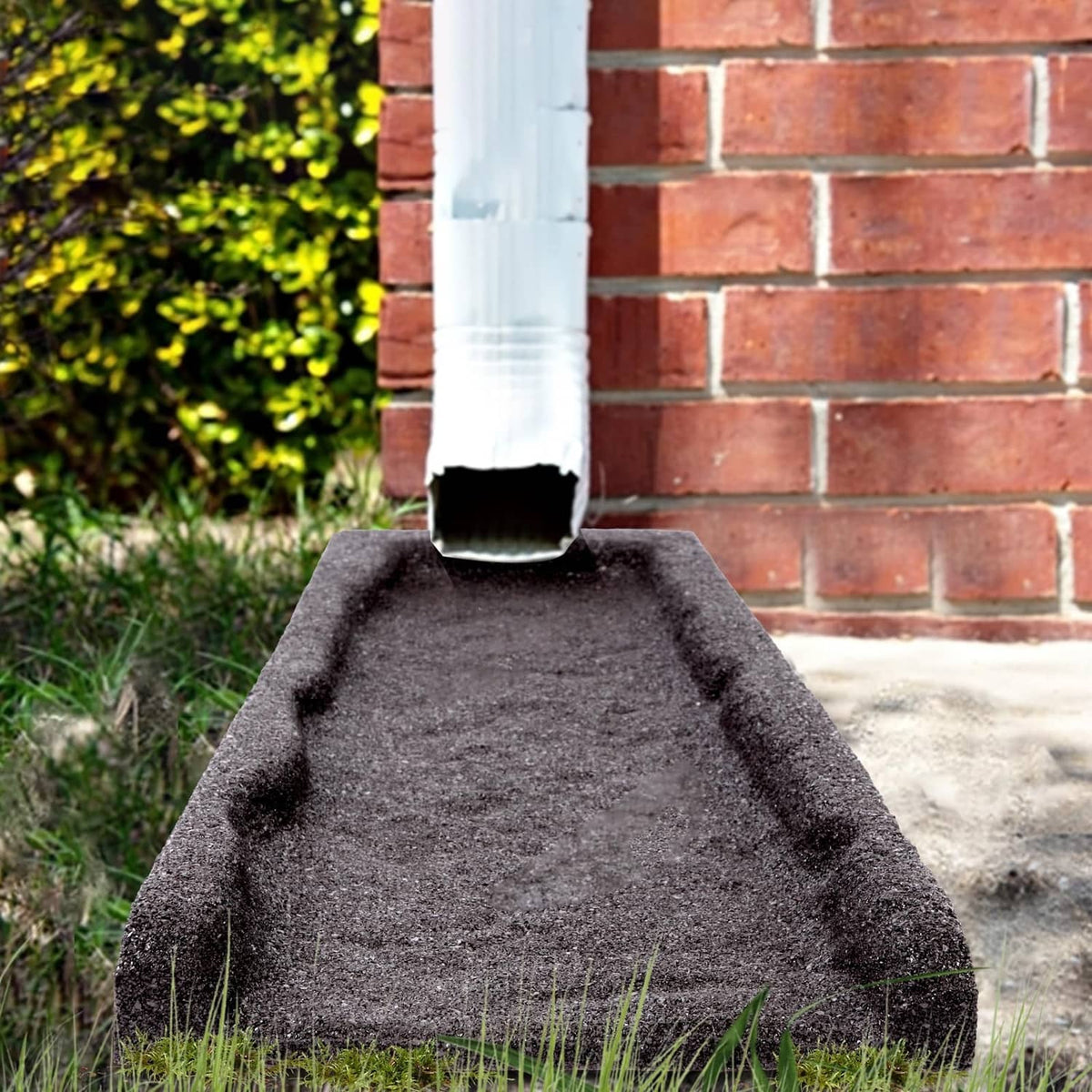 Brown rubber downspout splash block below a downspout pipe on a concrete path