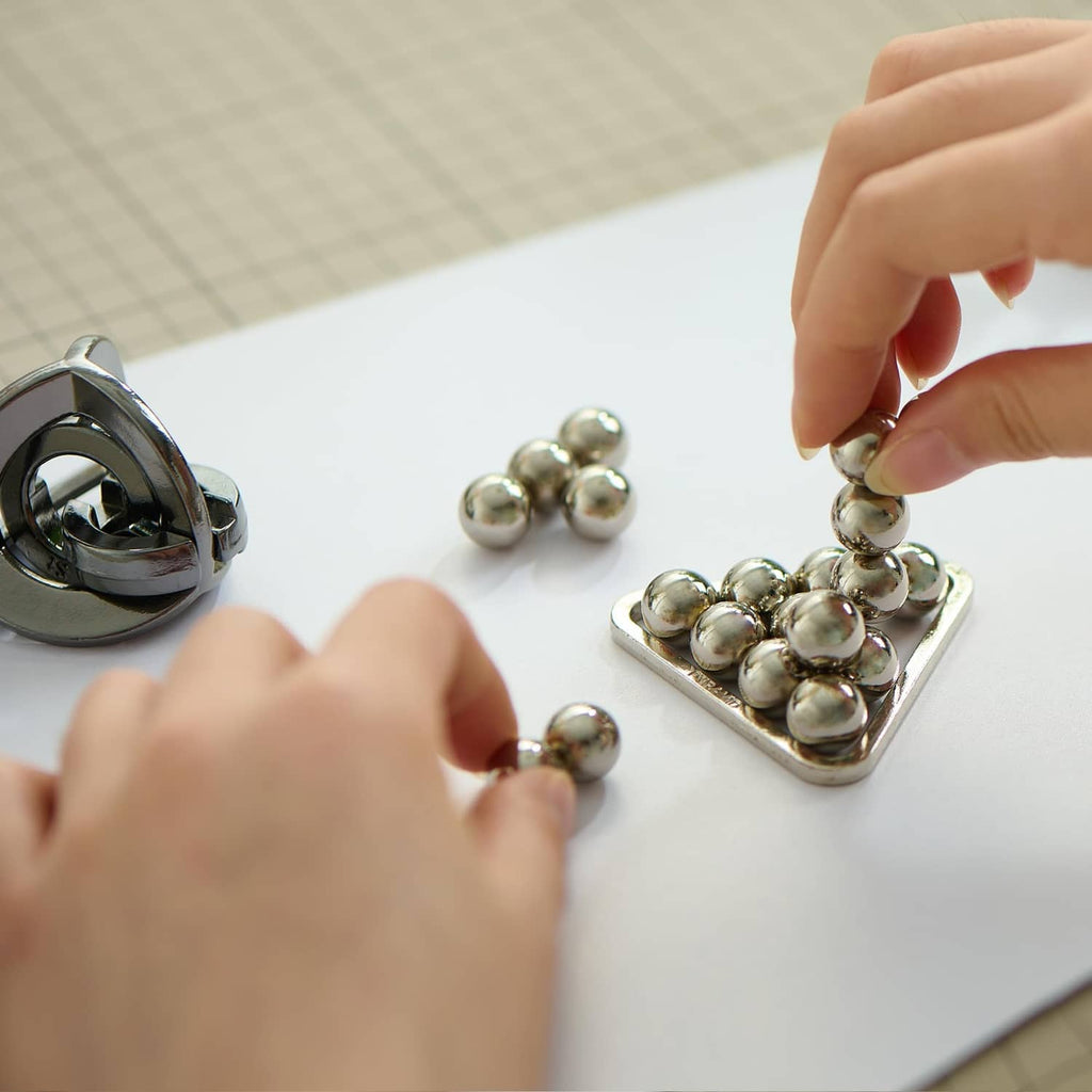 Hands assembling a silver-tone bead puzzle on tray, with triangular ring piece