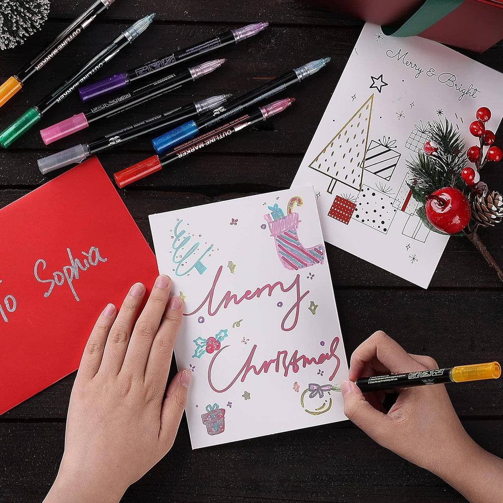 Metallic outline markers coloring cards with holiday designs on dark tabletop.
