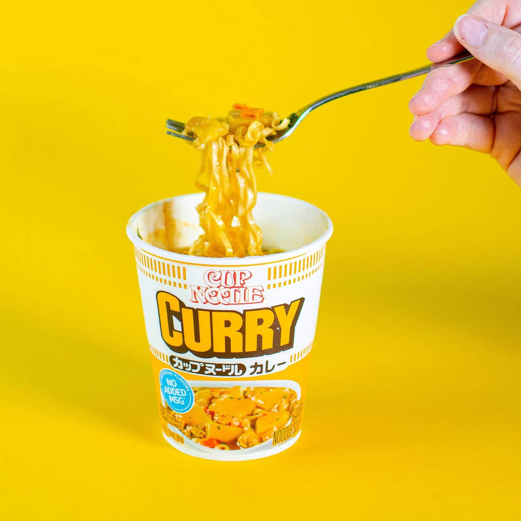 Fork lifting steaming noodles above curry cup noodles in white cup
