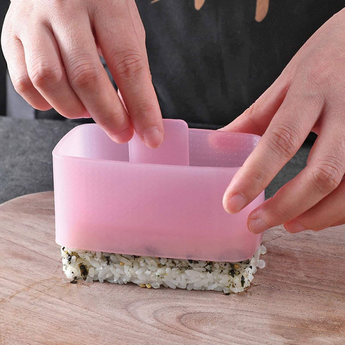 Hands pressing rice into a pink onigiri mold tray using the included plastic press