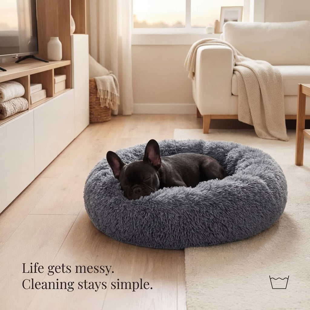 Dark grey round plush dog bed on floor with small dog curled up.