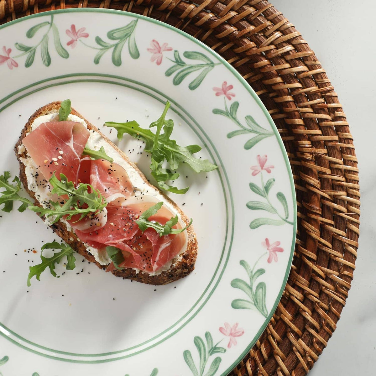 Close-up floral dinner plate with sliced cured meat, greens, and black pepper specks