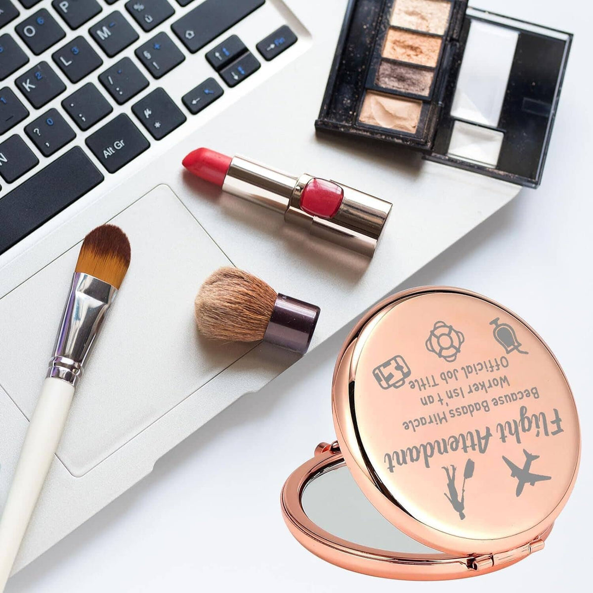 Rose-gold round makeup mirror compact with etched design, beside lipstick and brushes.