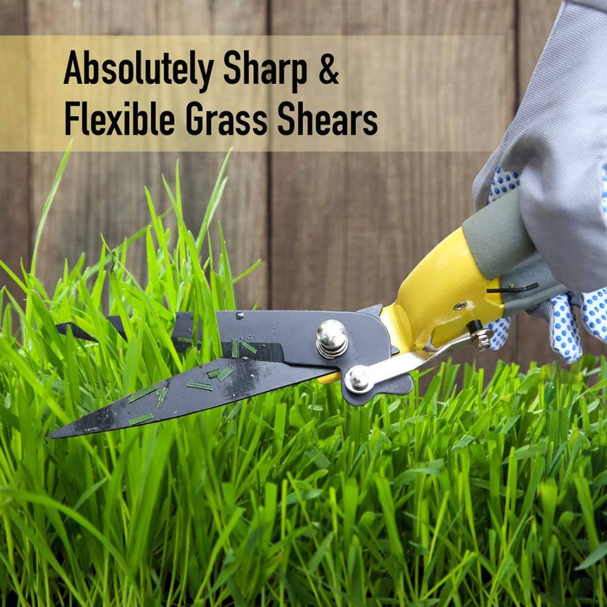 Grass shears with yellow handle and gray blades trimming lawn grass near a wooden fence.