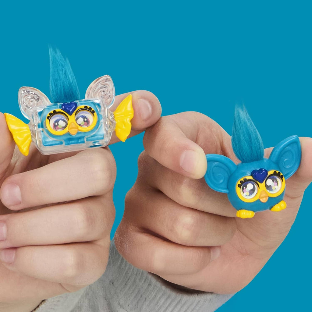 Hands holding two blue Furby minis toy figures with big yellow goggles and mohawks